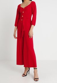 Mono rojo con escote cuadrado, mangas tres cuartos, botones de madera en la parte delantera y un cinturón a juego. Cuerpo ajustado con piernas amplias.