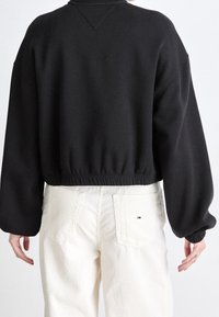 Felpa nera corta con colletto a girocollo e orlo elastico. Presenta maniche lunghe e larghe con polsini a coste. Indossata con pantaloni chiari.