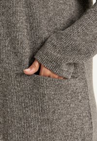 Grijze ribgebreide stof met een hand met vingers in een voorzak. De textuur is zacht, met afwisselend lichte en donkere garens.