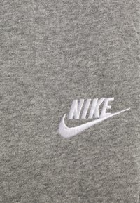 Grått tyg med vitt broderat Nike-logotyp och swoosh på en strukturerad textilbakgrund.