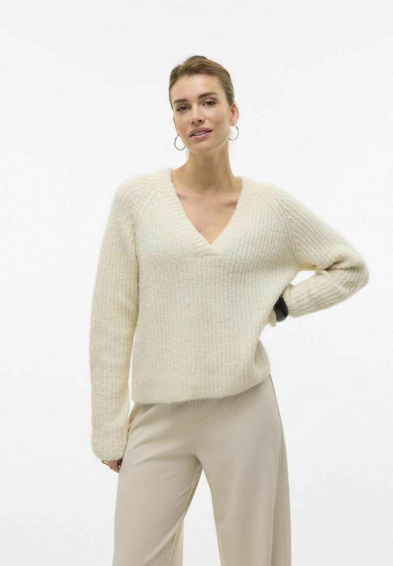 Pull en maille crème avec un col en V, des manches longues, un textile texturé et une coupe décontractée. Associé à un pantalon de couleur claire.