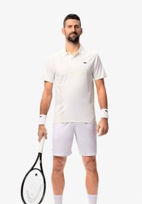 Hvid Lacoste polo med korte ærmer, parret med hvide shorts. Holder en sort tennisracket, og håndledsbind samt korte shorts fremhæver den atletiske stil.