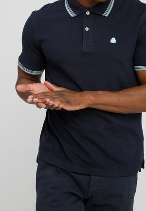 Uomo che indossa una polo blu navy con colletto e maniche a righe, si strofina le mani davanti a uno sfondo semplice.