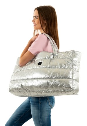 Jonge vrouw in een roze shirt en spijkerbroek draagt een grote zilveren gewatteerde draagtas over haar schouder, terwijl ze glimlacht naar de zijkant.