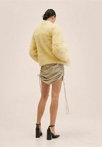 Kobieta z ciemnymi włosami upiętymi w kok, ubrana w żółtą puszystą kurtkę, beżową mini spódnicę z marszczeniami oraz czarne wysokie buty na obcasie z paskiem na kostce, stojąca tyłem.