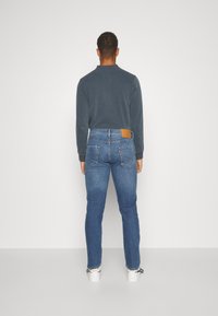 Vista trasera de una persona que lleva puestos unos jeans ajustados de mezclilla azul con un parche de cuero marrón claro en la parte trasera de la cintura, combinados con una camisa oscura de manga larga.