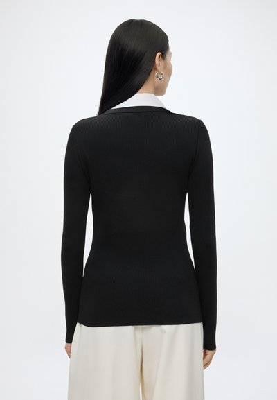 Femme aux longs cheveux noirs portant des boucles d'oreilles argentées en forme d'anneaux, un haut noir côtelé ajusté à manches longues et un pantalon blanc ample, vue de dos.