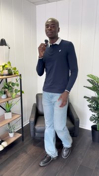 Tume sinise pika varrukaga polo särk valge kraega, koos helesiniste kõrge vöökoha teksapükstega ja mustade nööpidega kingadega. Siseruum, kus on taimi.