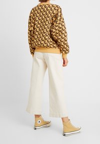 Persona vistiendo un suéter beige con estampado, pantalones anchos blancos y zapatillas Convers de caña alta amarillas, de pie contra un fondo blanco liso.