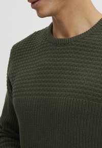 Dunkelgrüner Strickpullover mit einem strukturierten Design, ribbeltem Rundhalsausschnitt und einem glatten Unterteil. Weicher, mittelschwerer Stoff.