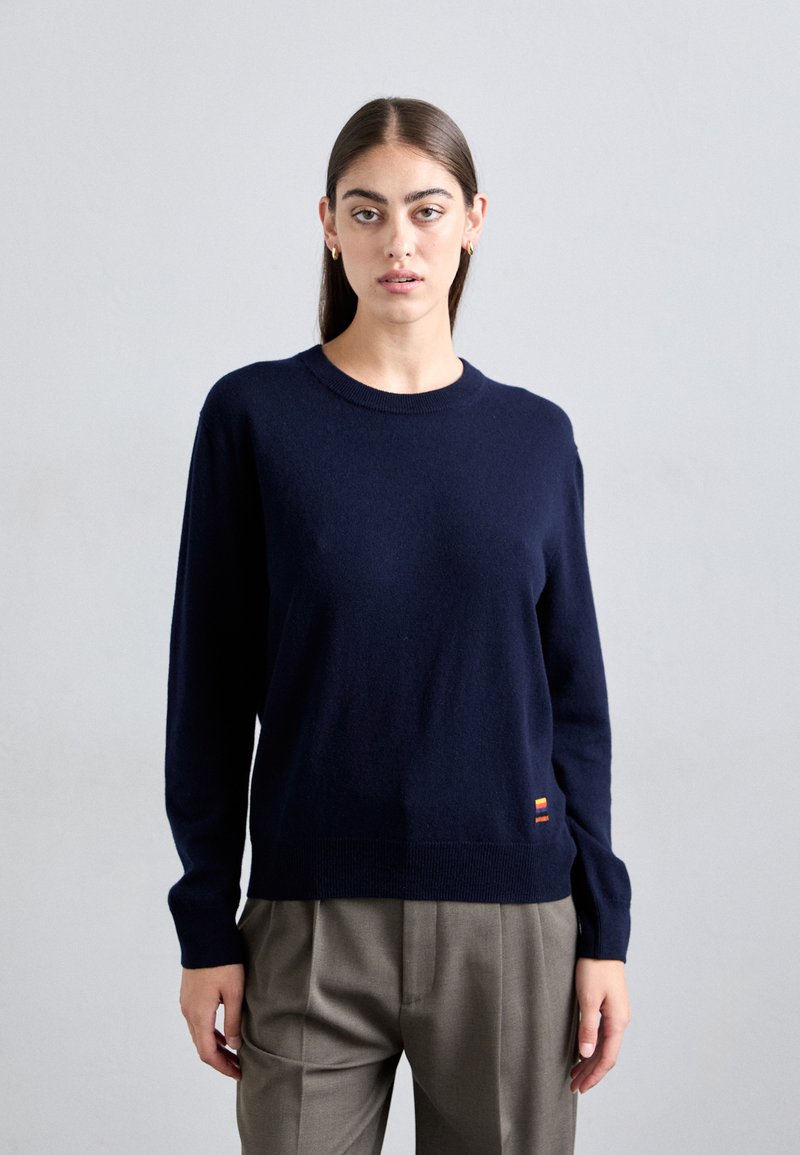 Navy blue knitted sweater with long sleeves, round neckline, and a ribbed hem. Features a small multicolored label at the lower left.