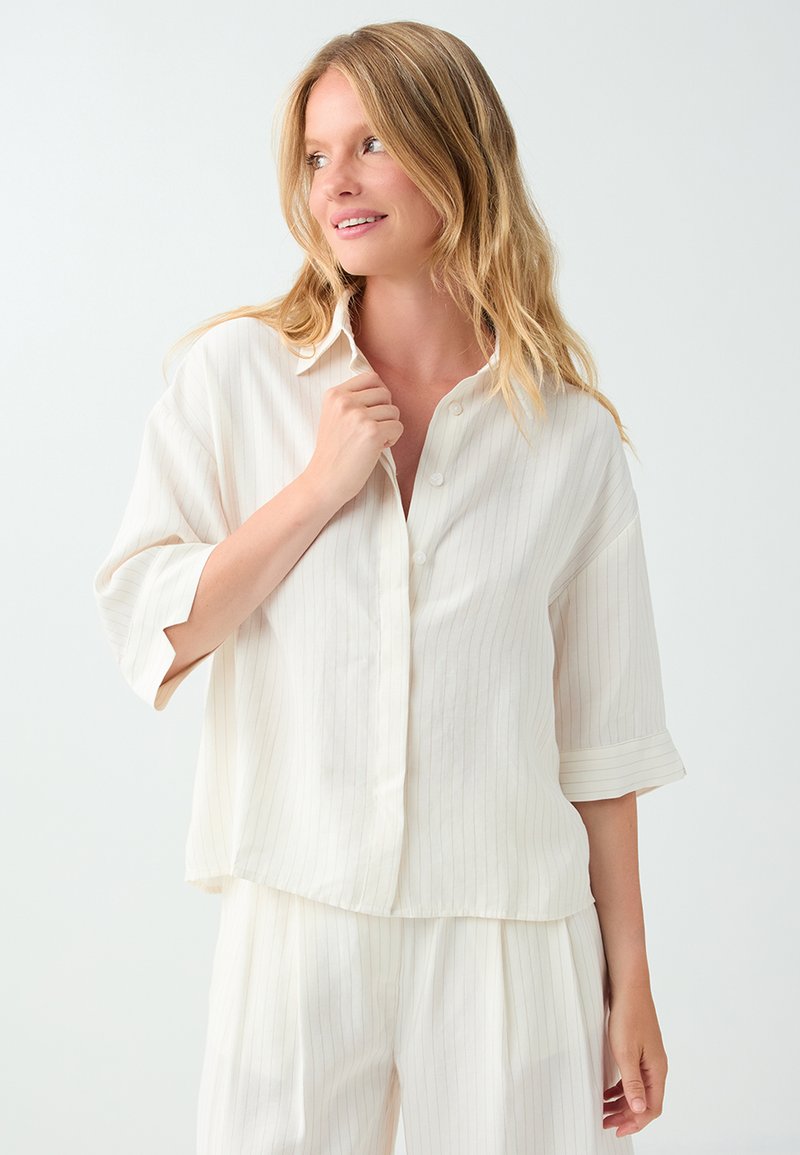 Femme aux longs cheveux blonds portant une chemise blanche ample à fines rayures boutonnée et un pantalon assorti, regardant sur le côté avec un léger sourire.