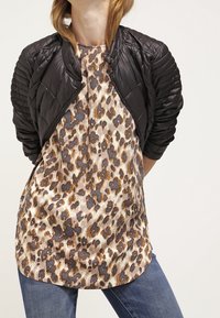 Giubbotto bomber trapuntato nero con collo alto, indossato sopra una maglia beige e marrone con stampa leopardata, abbinato a jeans in denim blu.