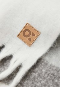 Merinowollen sjaal in het wit, voorzien van een tan kleurig leren label met een gegraveerd ontwerp. Zachte textuur, franjes aan de randen en een subtiel grijs patroon.