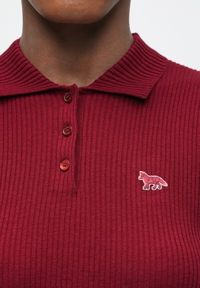 Tuvumā redzams cilvēks, kurš valkā ribotu tumši sarkanu polo kreklu ar trim pogām un mazu izšūtu lapsas logo uz krūtīm.