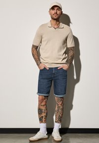 Camisa polo beige de manga corta, pantalones cortos de mezclilla azul con puños enrollados, zapatillas blancas y una gorra de color claro. Presenta tatuajes prominentes en las piernas.