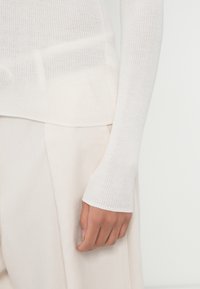 Camisola de mangas compridas com textura canelada em off-white, apresentando punhos ajustados e uma malha leve; combinada com calças de cor clara correspondentes.