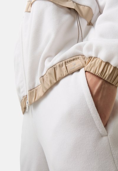 Bílá fleecová bunda s béžovými saténovými akcenty, se zipovým zapínáním, elastickými manžetami a boční kapsou. Texturovaná tkanina pro teplo.