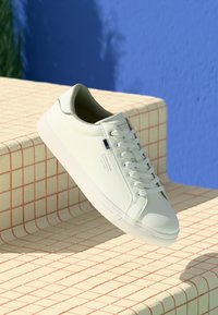 White low-top sneaker with laces on tiled stairs with red grid lines, blue wall background, and partial shadow visible.