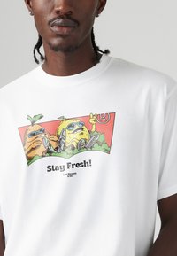 T-shirt bianco in cotone con una stampa grafica colorata che raffigura due frutti cartoon con occhiali da sole, con la scritta "Stay Fresh!" al centro.