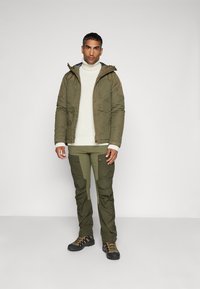 Olijfgroene geïsoleerde jas met een capuchon, een witte gebreide trui eronder, bijpassende olijfgroene cargobroek en zwarte wandelschoenen.