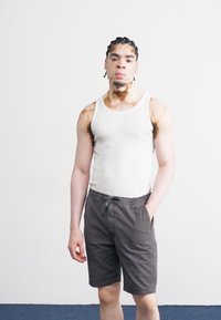 Débardeur blanc à côtes et short gris foncé. Le modèle a les cheveux tressés et se tient contre un fond uni, les mains dans les poches.