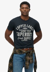 Marineblauw katoenen T-shirt met grote witte grafische tekst. Gecombineerd met lichtblauwe jeans en een geruit flanellen overhemd dat om de taille is gestrikt.