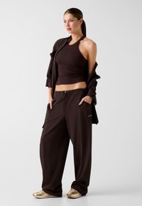 Débardeur marron côtelé associé à un pantalon marron ample, avec poches et cordon de serrage. Le mannequin porte des baskets beige.