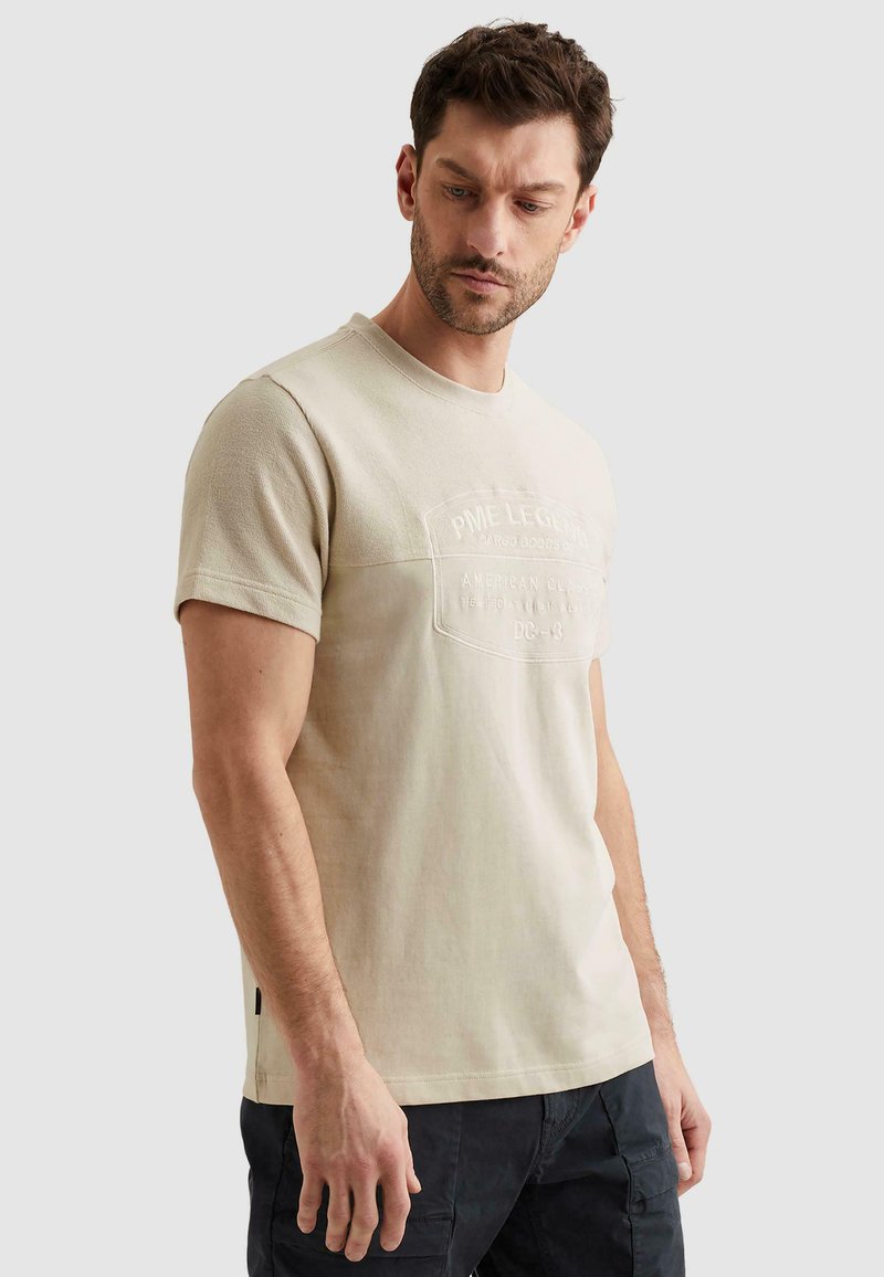 Homme aux cheveux courts portant un t-shirt beige à manches courtes avec un logo en relief, regardant vers le bas, debout devant un fond clair uni.