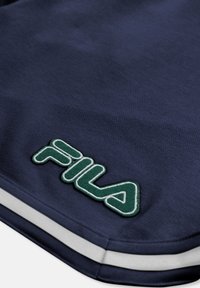 Gros plan sur un tissu bleu marine avec un patch logo FILA vert et blanc et une bande horizontale blanche près du bord.