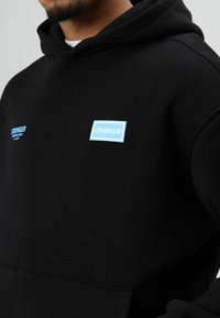 Sweat à capuche en coton noir avec des motifs bleus comprenant les mots "STRONGER" et "chance" sur la poitrine, coutures doubles, et une poche kangourou classique.