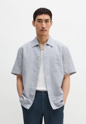 Mann trägt hellblau gestreiftes Kurzarmhemd über weißem T-Shirt mit dunkelblauer Hose, Hände in den Taschen, neutraler Hintergrund.