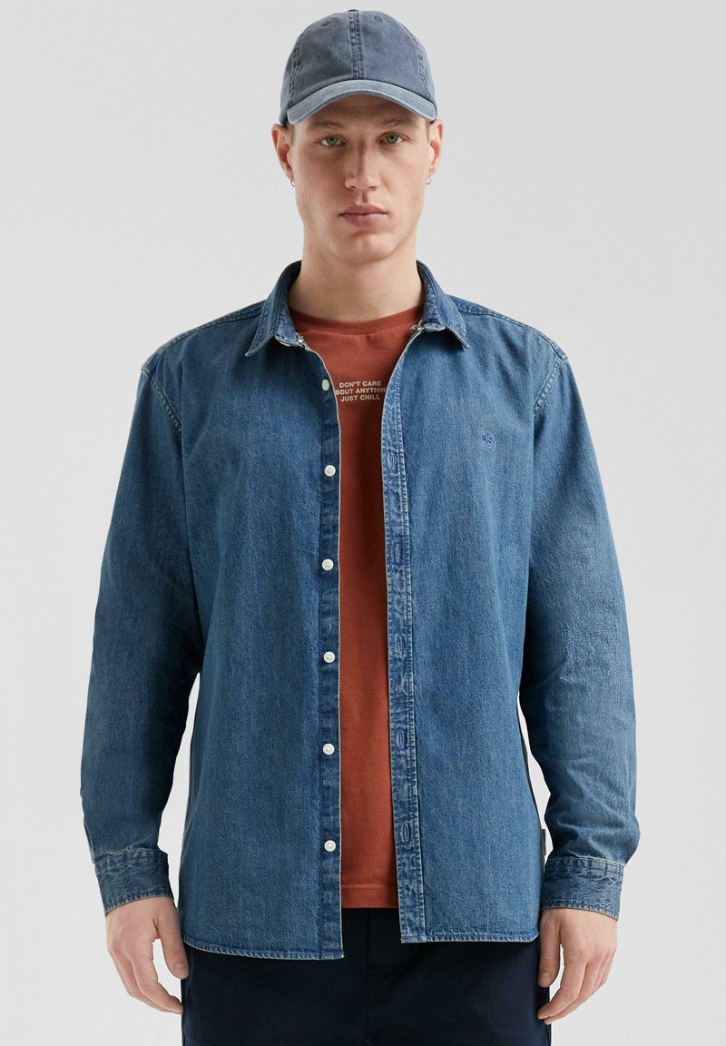 Denim shirt, medium wash, button-down style, met twee borstzakken, gecombineerd met een roestkleurig T-shirt en een blauwe baseballcap.
