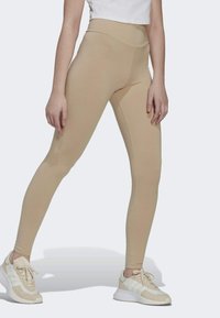 Frau in beigen High-Waist-Leggings und beigen-weißen Sneakers, die mit gespreizten Beinen vor einem schlichten Hintergrund steht.