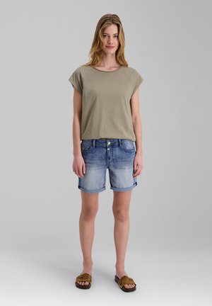 Woman standing wearing a khaki short-sleeve top, rolled denim shorts, and brown open-toe sandals against a plain gray background.