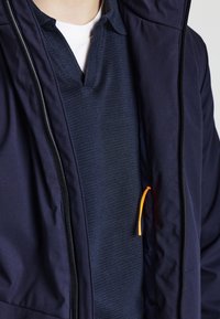 Veste bleu marine avec une fermeture éclair à l'avant, comportant un polo bleu marine texturé en dessous. Un cordon de serrage orange est visible.