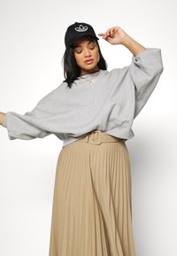 Felpa grigia oversize con maniche ampie abbinata a una gonna plissettata beige e un cappellino nero con logo, indossata con collane a strati.