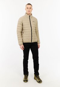Jonge man staat in een beige gewatteerde jas, zwarte broek en olijfgroene wandelschoenen tegen een effen witte achtergrond.