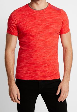 Man draagt een nauwsluitend rood gemêleerd T-shirt met korte mouwen, gecombineerd met een zwarte broek, staand tegen een effen achtergrond.