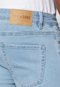 Los jeans de mezclilla azul claro presentan un parche de cuero etiquetado "ONLY & SONS" con costuras y un bolsillo trasero para mayor funcionalidad.