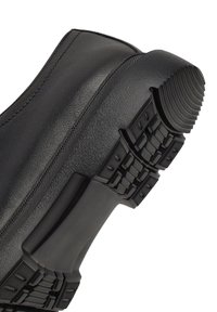 Scarpa in pelle nera con suola in gomma testurizzata caratterizzata da un ampio motivo di grip, bordi rinforzati e un profilo superiore liscio.