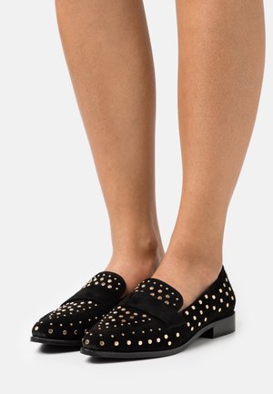 Musta mokkakengät, joissa on kultaiset niittiakcentit, pyöreä kärki ja tasapohja. Suunnittelussa on sileä nauha yläosan yli.