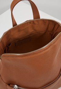 Intérieur d'un sac à dos en cuir marron avec une texture lisse ; fermeture éclair sur le dessus et une poche zippée sur le côté ; doublé de tissu marron.