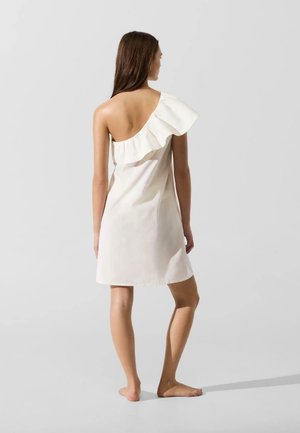 Kobieta stojąca boso, ubrana w białą sukienkę z odsłoniętym jednym ramieniem i marszczonym dekoltem, zwrócona tyłem na jednolitym jasnym tle.