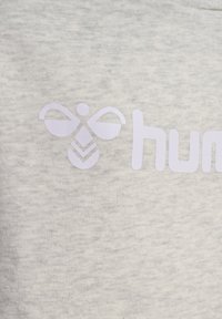 Hellgrauer Stoff mit einem weißen Logo einer Biene und dem Wort "hum" in einer modernen Schriftart. Die Textur wirkt weich und glatt.