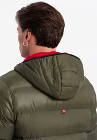 Olivgrön puffjacka med huva, med quiltad textur och en röd logotypaccent. Den inre kragen är röd med en svart kant.