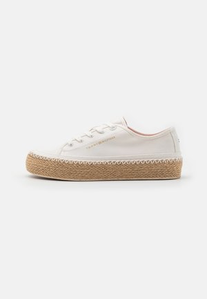 Zapatilla deportiva de lona blanca con suela plataforma tejida beige, cordones blancos y el logo dorado "Tommy Hilfiger" en el lateral, vista de perfil izquierdo.