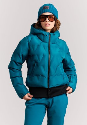Persoon draagt een teal donsjack met capuchon, bijpassende broek, blauwe gebreide muts en grote amberkleurige zonnebril tegen een effen achtergrond.