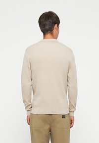 Beige gebreide pullover met lange mouwen en een kraag. Heeft een textuurpatroon en een rechte zoom. Ggedragen met khaki broeken.