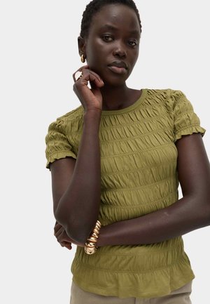 Donna con pelle scura che indossa una maglietta verde oliva arricciata a maniche corte, orecchini a cerchio dorati, un grande braccialetto dorato e un anello d'oro, in posa pensierosa.